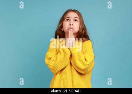 Portrait of Little girl schaut zu Gott beten mit flehenden Augen, so dass Kindheit Wunsch und Hoffnung, tragen gelben lässigen Stil Pullover. Innenaufnahme des Studios isoliert auf blauem Hintergrund. Stockfoto