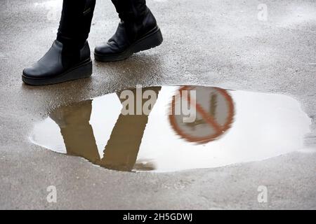 Regen in der Stadt, Frau in Stiefeln läuft um die Pfütze auf der Stadtstraße. Verkehrszeichen, die eine Abbiegung verbieten, spiegeln sich im Wasser wider, Konzept der falschen Wahl Stockfoto