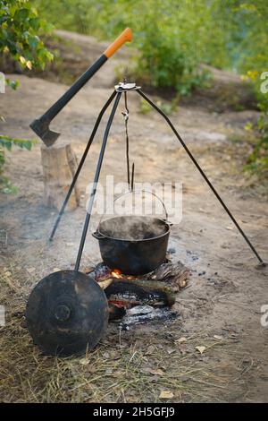 Ein Lagertopf, eine Axt, Feuerholz und ein Lagerfeuer. Zubereitung von Speisen am Lagerfeuer in wildem Camping, Ruhe in der Natur. Stockfoto