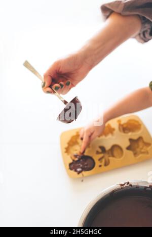 Kinder- und Erwachsenenhände gießen auf einem weißen Tisch eine Metalllöffel Schokoladenteig in eine Silikonform. Weihnachts-Lebkuchen. Stockfoto