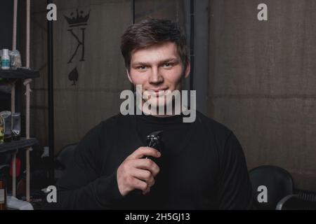 Ein zotteliger lächelnder Typ mit kaukasischem Aussehen und dunklem Haar hält eine elektrische Rasiermaschine und Haarschnitte in der Hand. Friseursalon. Stockfoto