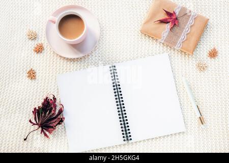 Leerer offener Notizblock, Kaffeetasse, Geschenk in einer Schachtel, Stift und Herbstblätter auf weißem gestricktem karierten Hintergrund. Draufsicht, flach liegend Stockfoto