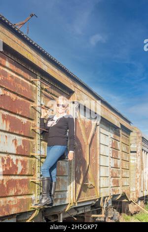 Porträt einer erwachsenen blonden Frau, die eine verlassene Zugwagenleiter klettert. Konzept für verlassene Orte Stockfoto