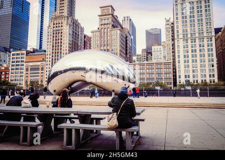 Menschen sitzen auf Bänken vor Sir Anish Kapoors Skulptur Cloud Gate, die den Spitznamen The Bean trägt, vor der Skyline von Chicago, IL, USA Stockfoto