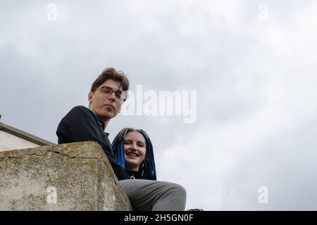 Ein Mann und eine Frau umarmen sich, sitzen auf einer Betonbrüstung. Die Frau ist lässig mit langen blauen Dreadlocks gekleidet. Mann in Geschäftskleidung. Blick vom Th Stockfoto