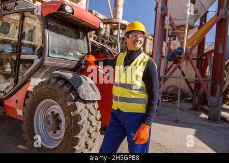 Fröhlicher männlicher Arbeiter, der in der Fabrik neben dem Traktor steht Stockfoto