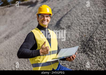 Fröhlicher Fabrikarbeiter, der Dokumente hält und Daumen hoch gibt Stockfoto