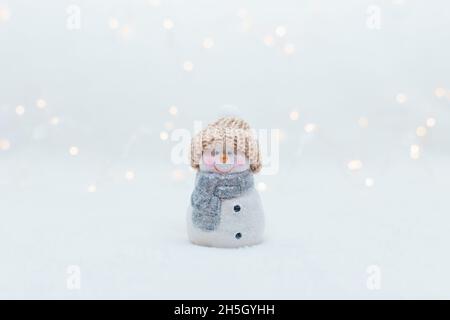 Dekorative Figuren im Weihnachtsstil. Die Statuette eines Schneemanns in einem gestrickten Hut auf weißem Hintergrund. Festliche Einrichtung, warme Bokeh-Lichter Stockfoto