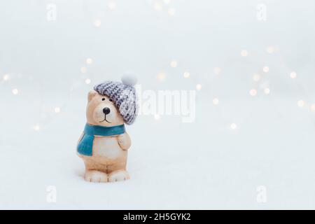 Dekorative Figuren im Weihnachtsstil. Die Statue eines Bären in einem gestrickten Hut auf weißem Grund. Festliche Einrichtung, warme Bokeh-Lichter Stockfoto