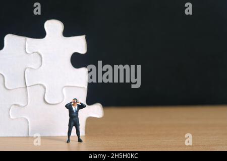 Geschäftsleute, die vor einem Puzzle-Puzzleteil stehen, stapeln mit einem Fernglas auf einem Holztisch. Miniatur kleine Menschen Spielzeug Fotografie. Stockfoto