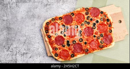 Rechteckige Pizza mit Pepperoni, Käse und Oliven auf einem rechteckigen Holzbrett auf dunkelgrauem Hintergrund. Draufsicht, flach liegend Stockfoto