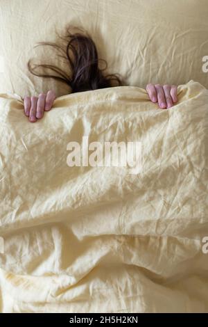 Das Mädchen im Bett ist mit einer gelben Decke mit dem Kopf bedeckt, man sieht einen Teil ihrer Hände und Haare. Das Konzept, sich vor Herbst und Winter zu verstecken. Stockfoto