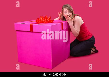 Nahaufnahme einer jungen hübschen Dame in rotem T-Shirt und schwarzer Jeans, die auf dem Boden neben der großen, pinken Schachtel sitzt und ihren Mund bedeckt Stockfoto