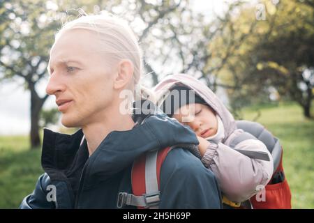 Mann, der mit einem verklenkten Kleinkind in den Berg wandert. Kind sitzt im Rucksack auf dem Rücken des Vaters. Aktives Familienabenteuer im Wald mit müdem Mädchen. Kalt Stockfoto