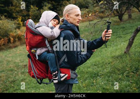 Vater fotografiert beim Wandern mit Kind im Rucksack. Vlogger-Mann, der mit einem erlafften Kind im Rucksack in den kalten Bergen läuft. Aktive Zeit mit Stockfoto