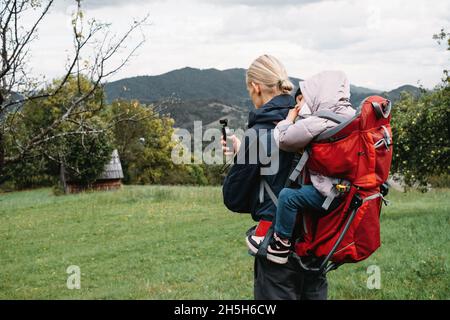 Vater fotografiert beim Wandern mit Kind im Rucksack. Vlogger-Mann, der mit einem erlafften Kind im Rucksack in den kalten Bergen läuft. Aktive Zeit mit Stockfoto