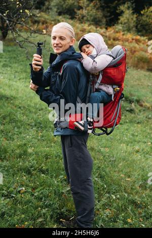 Vater fotografiert beim Wandern mit Kind im Rucksack. Vlogger-Mann, der mit einem erlafften Kind im Rucksack in den kalten Bergen läuft. Aktive Zeit mit Stockfoto