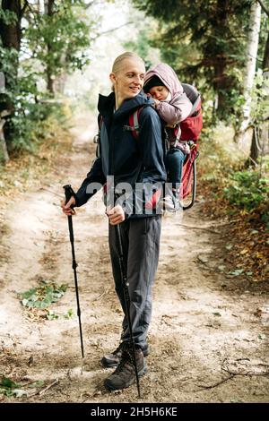 Mann, der mit einem verklenkten Kleinkind in den Berg wandert. Kind sitzt im Rucksack auf dem Rücken des Vaters. Aktives Familienabenteuer im Wald mit müdem Mädchen. Kalt Stockfoto