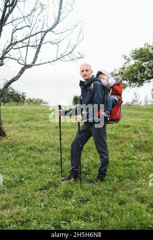 Mann, der mit einem verklenkten Kleinkind in den Berg wandert. Kind sitzt im Rucksack auf dem Rücken des Vaters. Aktives Familienabenteuer im Wald mit müdem Mädchen. Kalt Stockfoto