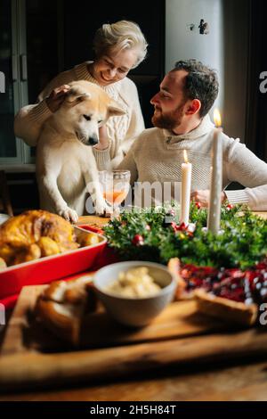 Vertikale Aufnahme eines glücklichen jungen verliebten Paares, das während der Weihnachtsfeier mit dem Hund Akita Inu am festlichen Weihnachtstisch sitzt. Stockfoto