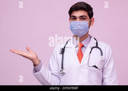 Junger Arzt trägt Maske hält etwas in leeren Hand isoliert Hintergrund. Stockfoto