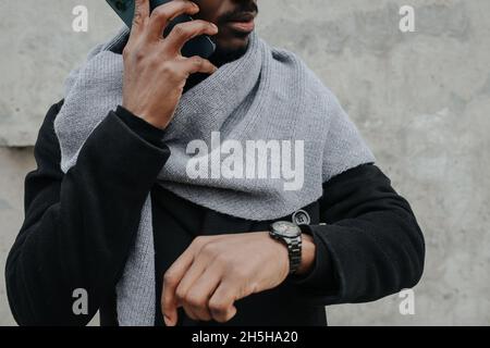 Zugeschnittenes Bild eines beschäftigten schwarzen Mannes im Freien, der am Telefon telefoniert, während er die Zeit überprüft. Er ist warm gekleidet in einen Mantel und Schal. Stockfoto