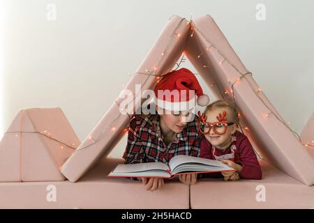 Mutter mit Tochter im Schlafanzug, Weihnachtsmütze und Stirnband beim Lesen eines Buches in einem Babyzimmer. Nahaufnahme einer jungen Frau und ihres Mädchens, in dem sie sich entspannen Stockfoto