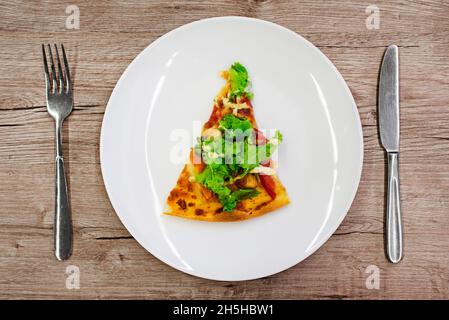 Eine Scheibe Pizza auf einem weißen Teller, eine Gabel, ein Messer, eine Pizza auf einem Holztisch. Stockfoto