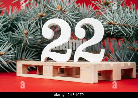 Frohes Neues Jahr 2022. Weiße Holznummern 22 stehen mit saftig grünen Christbaumzweigen auf Europalette auf rotem Hintergrund. Frohe Weihnachten. Stockfoto