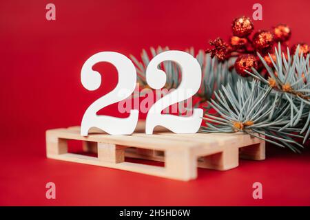 Frohes Neues Jahr 2022. Weiße Holznummern 22 stehen mit üppig grünem Christbaumzweig und Beeren auf Europalette auf rotem Hintergrund. Frohe Weihnachten Stockfoto