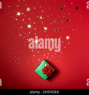Glänzendes grünes kleines Geschenk auf rotem Hintergrund mit funkelnden goldenen Sternen. Festliche Kulisse für Weihnachten, Neujahr. Flach liegend Stockfoto