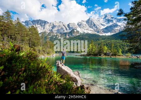 Junger Mann steht auf einem Felsen, Blick in die Ferne, Eibsee und Zugspitze im Frühjahr mit Schnee, Wettersteingebirge, bei Grainau, Obere Stockfoto