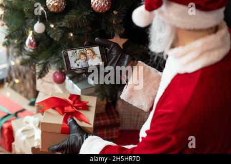 Über der Schulter sehen Sie, wie der Weihnachtsmann, der Weihnachtsgeschenk und Smartphone hält, während er Geschenke von zwei Kindern stiehlt Stockfoto