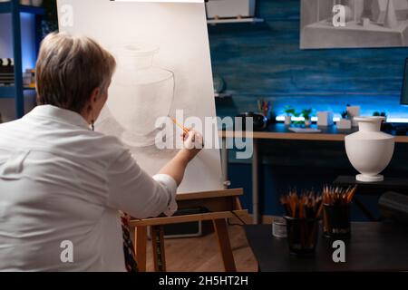 Kaukasisch alter Künstler, der mit Vase und Bleistift auf Leinwand für authentisches Zeichnen in der Werkstatt arbeitet. Ältere Frau mit künstlerischem Hobby Meisterwerk für Bildende Kunst-Projekt im Studio zu schaffen Stockfoto