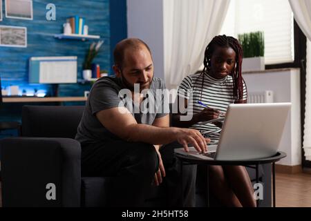 Junges interrassisches Paar, das zuhause Steuern für die Finanzwirtschaft zahlt. Multiethnische Partner, die mit einem Laptop und einem Notebook Steuerzahlungen leisten und mit Technologie Budgetgelder berechnen Stockfoto