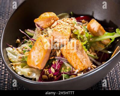 Grünen Salat mit gebratenem Lachsfilet und Nüssen in dunkler Schüssel auf schwarzer Korbplatte mischen Stockfoto