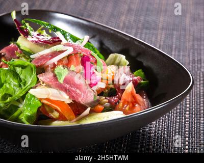 Salat mit Rinderscheiben, grünem frischen Salat, Gurken, Tomaten und Zwiebeln in einer Schüssel auf schwarzer Korbplatte Stockfoto