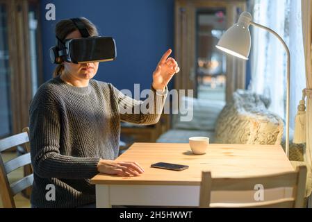 Frau in Strickpullover mit vr Virtual Reality-Headset im Wohnzimmer. Für Arbeit oder Unterhaltung Stockfoto
