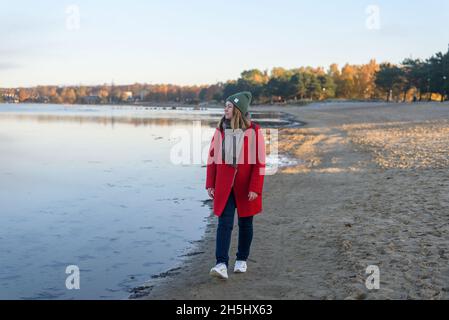 Frau im roten Mantel, die am kalten Morgen bei Sonnenaufgang in der Nähe des Meeres spazieren ging Stockfoto