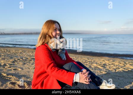 Frau in rotem Mantel sitzt in der Nähe des Meeres bei Sonnenaufgang kalten Morgen Stockfoto