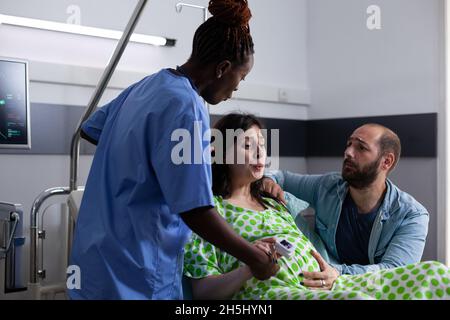 Medizinischer Assistent, der die Hand einer Schwangeren hält, die im Bett der Krankenstation sitzt. Der junge Mann unterstützt die Frau, die sich auf die Arbeit und die Geburt in der medizinischen Klinik vorbereitet, während die Krankenschwester assistiert Stockfoto