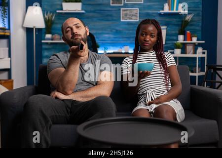 POV von interracial Menschen Fernsehen und entspannen zu Hause, Blick auf die Kamera. Kaukasischer Mann mit TV-Fernbedienung zum Umschalten der Kanäle und schwarze Frau mit Popcorn Stockfoto