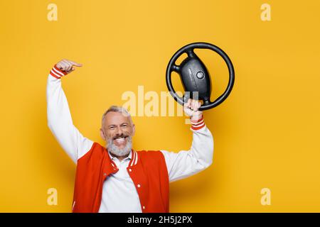Glücklicher Mann mittleren Alters in Bomberjacke, der auf gelbes Lenkrad zeigt Stockfoto