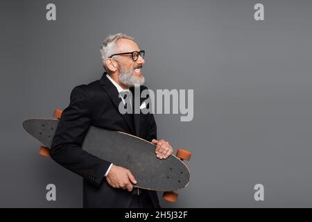 Fröhlicher Geschäftsmann mittleren Alters in Anzug und Brille mit Longboard auf Grau Stockfoto