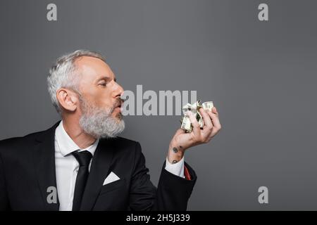 Seitenansicht eines bärtigen Geschäftsmannes mittleren Alters, der auf zerknitterte Dollar-Banknoten bläst, die isoliert auf Grau liegen Stockfoto