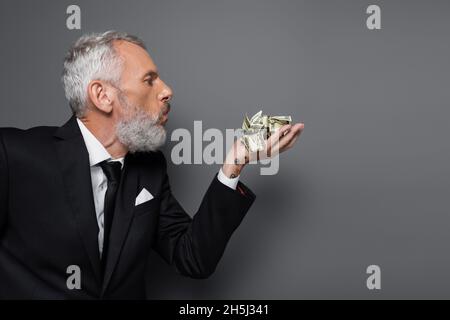 Seitenansicht eines tätowierten Geschäftsmannes mittleren Alters, der auf zerknitterte Dollarnoten auf Grau bläst Stockfoto