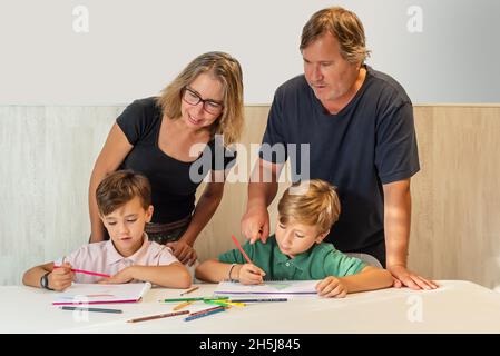 Zwei Kinder machen ihre Hausaufgaben zu Hause, während ihre Eltern ihnen zuschauen und ihnen helfen Stockfoto