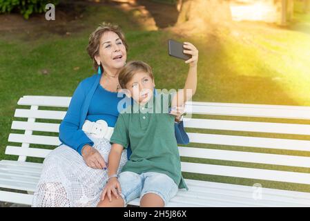 Enkel und Großmutter machen ein Selfie mit einem Handy, das auf einer Parkbank sitzt. Konzept von alten Menschen und neuen Technologien Stockfoto
