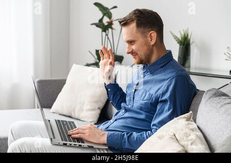 Ein lächelnder junger Mann sitzt auf der bequemen Couch im Wohnzimmer, entspannt sich und spricht mit seinem Freund oder seiner Familie, winkt am Laptop-Bildschirm und bleibt während einer Pandemie in Verbindung Stockfoto
