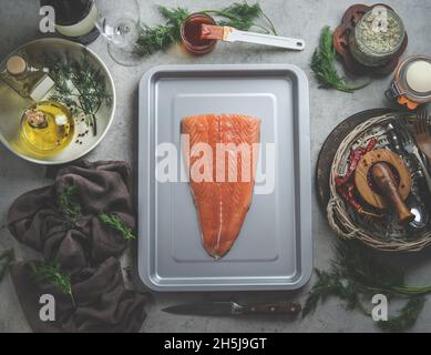 Rohes Lachsfilet auf grauem Backblech am Küchentisch mit Kräutern und Gewürzen, Öl, Pinsel mit Marinade, Geschirrtuch am grauen Küchentisch. Marinade fis Stockfoto